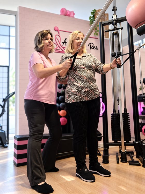Ana Carla Reis orientando uma aluna em exercício com aparelho de musculação